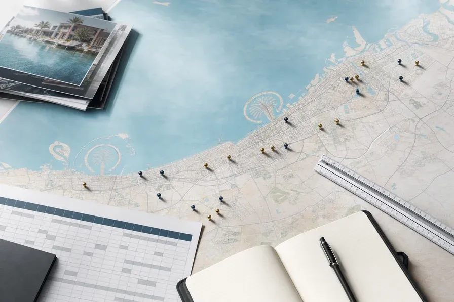 Overhead view of a UAE map spread across an architectural desk with gold and blue location pins marking various off-plan property communities, surrounded by brochures, a ruler, and a notebook.