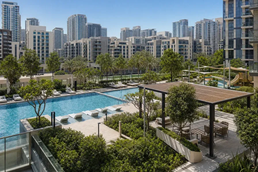 A modern Dubai residential tower podium level showing a landscaped deck with a swimming pool, shaded seating and greenery, set against a dense built-up urban district of mid-rise and high-rise apartments