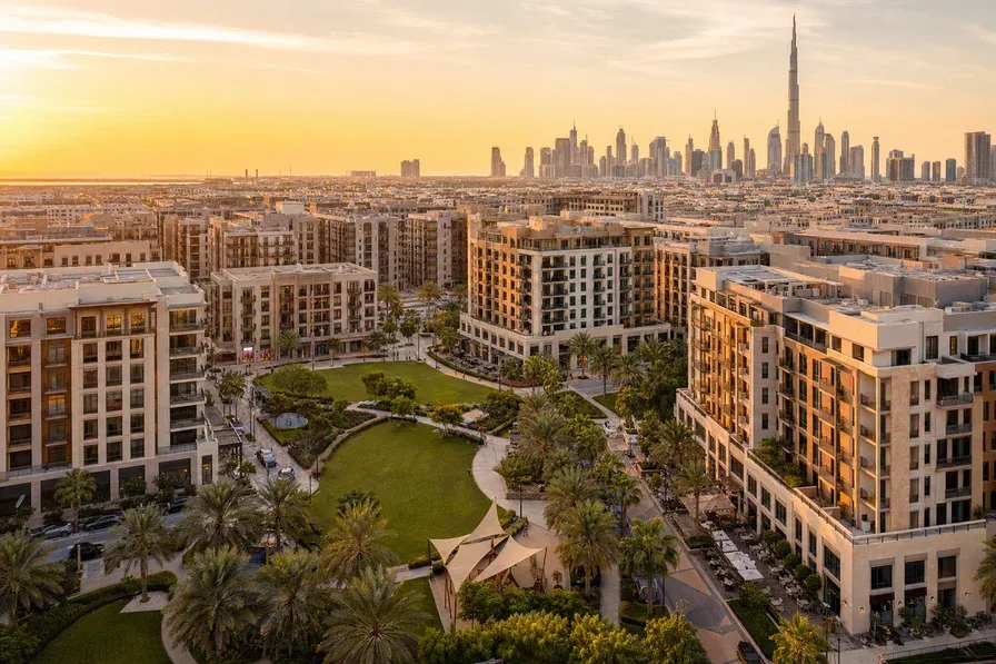 A wide-angle aerial photograph of a Dubai apartment-led community at golden hour, mid-rise residential towers clustered around a central park, palm-lined streets and ground-floor retail visible.